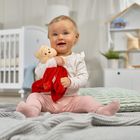 Ein lachendes Baby sitzt auf einer Decke und hält ein Kuscheltier. Im Hintergrund steht ein weißes Kinderbett.