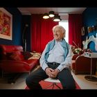 Ein älterer Mann sitzt lächelnd in einem modernen Wohnzimmer mit rotem Sofa, bunten Vorhängen und einem CD-Regal.