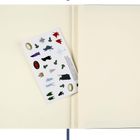 Ein Notizbuch mit weißen Seiten und einen Bogen mit bunten Illustrationen von Charakteren, Symbolen und Objekten dazwischen.