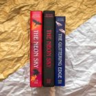 Drei Bücher mit den Titeln: "THE NEON SKY", "THE NEON SKY", "THE GLITTERING EDGE" vor golden-silbernem Hintergrund.