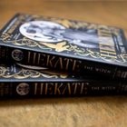 Zwei Bücher mit dem Titel "HEKATE THE WITCH" auf einem Holztisch. Goldene Verzierungen und eine Eule auf dem Cover.