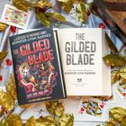 Zwei Bücher von Jennifer Lynn Barnes, Titel: "The Gilded Blade". Umgeben von Spielkarten und goldenen Blättern.