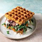 Zwei goldbraune Waffeln mit knusprigem Belag, grünem Gemüse und Soße, auf einem hellen Teller serviert.