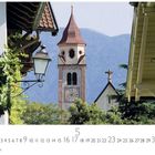 "Dorf Tirol", Kalenderblatt für Mai. Kirche mit Uhrturm vor Bergkulisse, umgeben von Bäumen und Gebäuden.