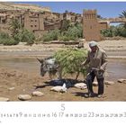 "Ait Benhaddou". Ein Mann führt einen Esel mit Grasbündel durch einen flachen Fluss, im Hintergrund Lehmhäuser und Palmen.