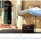 Text: "Caféhaus Nikosia". Eine alte, verwitterte Hausfassade mit grüner Tür, daneben ein Tisch mit Stühlen unter einem Sonnenschirm.