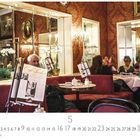 "Café Sacher" steht auf einem Zeitungshalter. Menschen sitzen an Tischen mit roten Bänken in einem elegant dekorierten Raum.