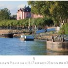 "WILLKOMMEN AN BORD." Im Hintergrund ein historisches, rosafarbenes Gebäude, gesäumt von Bäumen am Wasser.