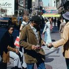 Text: "EMBASSY", "Salsa", "ONE WAY". Eine belebte Straßenkreuzung in einer Stadt mit mehreren Passanten und Verkehr.