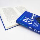 Der Titel "DA CAPO!" und der Name "Roland Zag" sind groß auf einem blauen Buchcover zu sehen. Öffnetes Buch daneben.