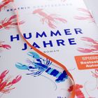 Titel: „Hummer Jahre“, Roman von Beatrix Gerstberger. Rote und blaue Hummer illustrieren das Design, orangefarbenes Band.