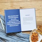 Ein Buch auf Holzuntergrund: Links Text "EIN SONG IST MANCHMAL ALLES...", rechts "Challenge 1". Daneben ein Teller mit Snacks.