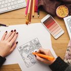 "ART BOOK" steht auf einem Regal. Eine Hand koloriert eine Illustration. Farbstifte und ein Smartphone sind sichtbar.