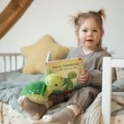 Ein Kind sitzt auf einem Bett mit einem Buch "Kleine Schildkröte, erzähl mir von dir" und einem Plüschtier-Schildkröte.