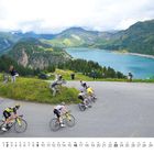 Kalender: Monat Mai unten. Radfahrer auf Bergstraße am See, umgeben von Bergen und Wäldern. Zuschauer am Straßenrand.