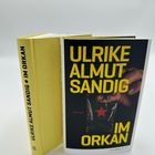 Zwei Bücher: „ULRIKE ALMUT SANDIG IM ORKAN“. Eines liegt, eines steht mit Cover: Hände, roter Stern, schwarze Hintergrund.