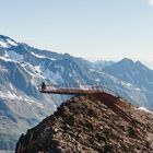 Zwei Personen stehen auf einer Metallplattform, die über eine felsige Berglandschaft hinausragt, umgeben von schneebedeckten Gipfeln.