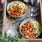 "The Good" auf Zeitungspapier. Zwei Schalen mit Nudeln, Karotten, Paprika und Fleisch, umgeben von Gewürzgefäßen und Tomaten.