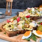 Ein Holzbrett mit belegtem Brot, Eierhälften und Gemüse. Im Hintergrund eine Sanduhr und Herbstdeko.