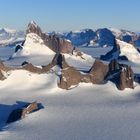 Verschneite, schroffe, felsige Berggipfel ragen aus einer weiten, eisbedeckten Landschaft empor.