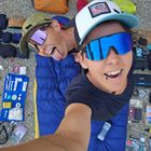 Zwei Personen mit Sonnenbrillen, umgeben von Camping- und Reiseutensilien, lächelnd und posierend.
