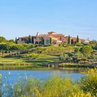 Kein Text erkennbar. Eine mediterrane Villa auf einem Hügel, umgeben von Bäumen und einem See im Vordergrund.