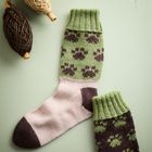 Zwei Socken mit grün-braunem Pfotenmuster, daneben drei Wollknäuel in Naturtönen auf einem grünen Hintergrund.