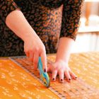 Eine Hand benutzt einen Rollschneider auf orangefarbenem Stoff mit weißen Mustern, daneben ein transparenter Lineal.