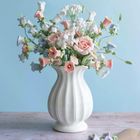 Gehäkelte Blumen in Pastellfarben stehen in einer weißen Vase auf einem Tisch; daneben Schere und Bindfaden.