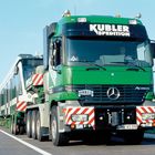 Aufschrift "KUBLER SPEDITION" auf einem grünen LKW, der ein großes Fahrzeug transportiert.
