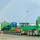 Ein großer, grüner Lastwagen mit der Aufschrift "Kübler Spedition" transportiert eine massive Maschine, im Hintergrund ein Regenbogen.