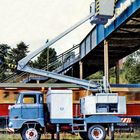 Man sieht ein hellblaues Nutzfahrzeug mit Hebebühne, das vor einer Überführung parkt. Im Hintergrund fährt ein roter Zug.
