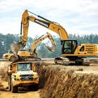 Ein Bagger mit der Aufschrift "AMAND" lädt Erde in einen Volvo-Kipper. Ein weiterer Bagger im Hintergrund. Wald im Hintergrund.