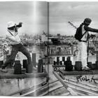 Texte: "Rolk". Zwei Personen auf einem Dach fotografieren in einer städtischen Umgebung. Schwarz-Weiß-Fotografie.