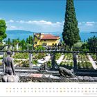 Mai-Kalenderseite: Ein schöner Rosengarten mit Skulpturen, Blick auf einen See, ein gelbes Gebäude und hohe Zypressen.