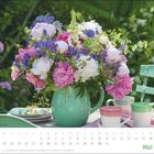 Kalendarium zeigt Mai. Strauß aus rosa, weißen und lila Blumen in grüner Vase auf Tisch mit Tassen.