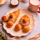Fröhliche Weihnachten. Zwei bärenförmige Brötchen auf einem Teller mit Zuckerstreuseln, Tasse Milch, kleines Deko-Nikolaus-Hütchen.