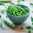 Eine Schale voller grüner Erbsen, umgeben von Erbsenschoten und Blättern auf einem Holztisch.