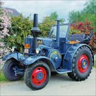 Text: "LANZ BULLDOG". Ein dunkelblauer Oldtimer-Traktor mit roten Felgen, stehend vor blühenden Büschen.