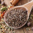 Ein Holzlöffel voller Kümmelsamen auf einem Tisch, umgeben von verschiedenen Gewürzen wie Kardamom, Zimt und Pfeffer.