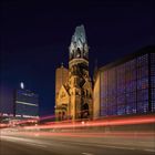 Ein nachts beleuchteter Turm mit beschädigter Spitze, daneben moderne Gebäude. Lichter ziehen horizontale Linien.