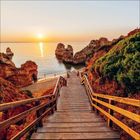 Ein hölzerner Steg führt zu einem Sandstrand, umgeben von felsigen Klippen, mit Sonnenuntergang über dem ruhigen Meer.