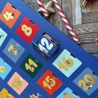 Ein Adventskalender mit Zahlen von 1 bis 24, bunte Karten, einige mit kleinen Tierillustrationen, Schokoladenstäbe im Hintergrund.