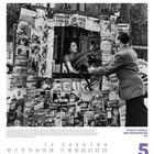 ROBERTO DOISNEAU DER ZEITUNGSKIOSK 1951. Schwarz-Weiß-Foto von einem Kiosk mit vielen Zeitschriften. Ein Mann hält einen Stuhl.