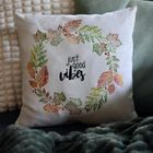 Text: "just good vibes". Ein Kissen mit herbstlichem Blätterkranz-Design, umgeben von weiteren Kissen und einer Decke.