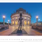 Das Bild zeigt das Bode-Museum in Berlin bei Dämmerung mit einer Statue im Vordergrund und erleuchteten Lampen entlang des Weges.