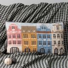 Ein Kissen mit gestricktem Muster bunter Häuser liegt auf einer schwarz-weiß gemusterten Decke, daneben zwei Wollknäuel.