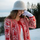 Eine lächelnde Person trägt eine rote, gemusterte Strickjacke und einen weißen Hut in winterlicher Landschaft.