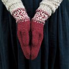 Rote, gemusterte Fäustlinge in betender Pose, getragen über grob gestrickten, beigefarbenen Ärmeln. Darunter ein dunkler Stoff.