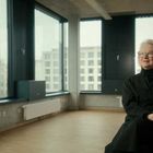 Ich kann keine Texte oder Logos erkennen. Eine ältere Person sitzt in einem modernen Raum mit großen Fenstern.